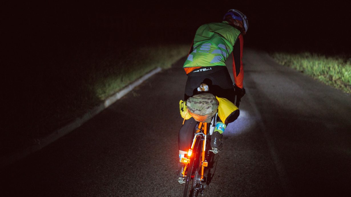 Cap number 101 of the #TPRNo1 ultracycling race riding away from camera into the dark on a rural road